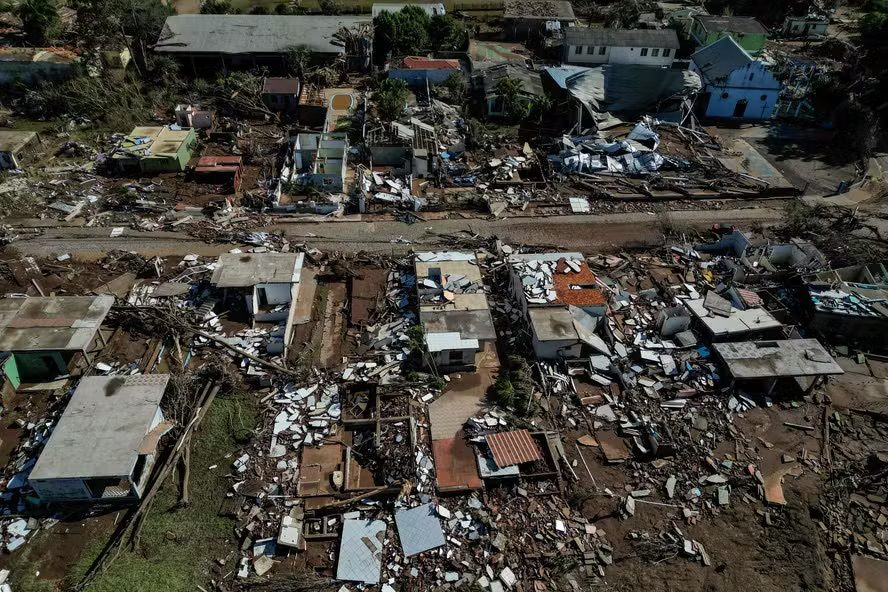 Vista aérea de bairro devastado após enchente, com casas destruídas, telhados arrancados e escombros espalhados pelas ruas.